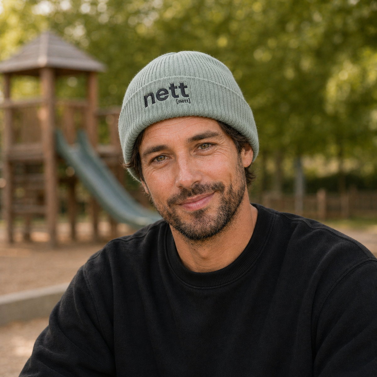 Mann vor einem Spielplatz trägt die JoisPulse Fisherman Beanie mit dem Statement nett