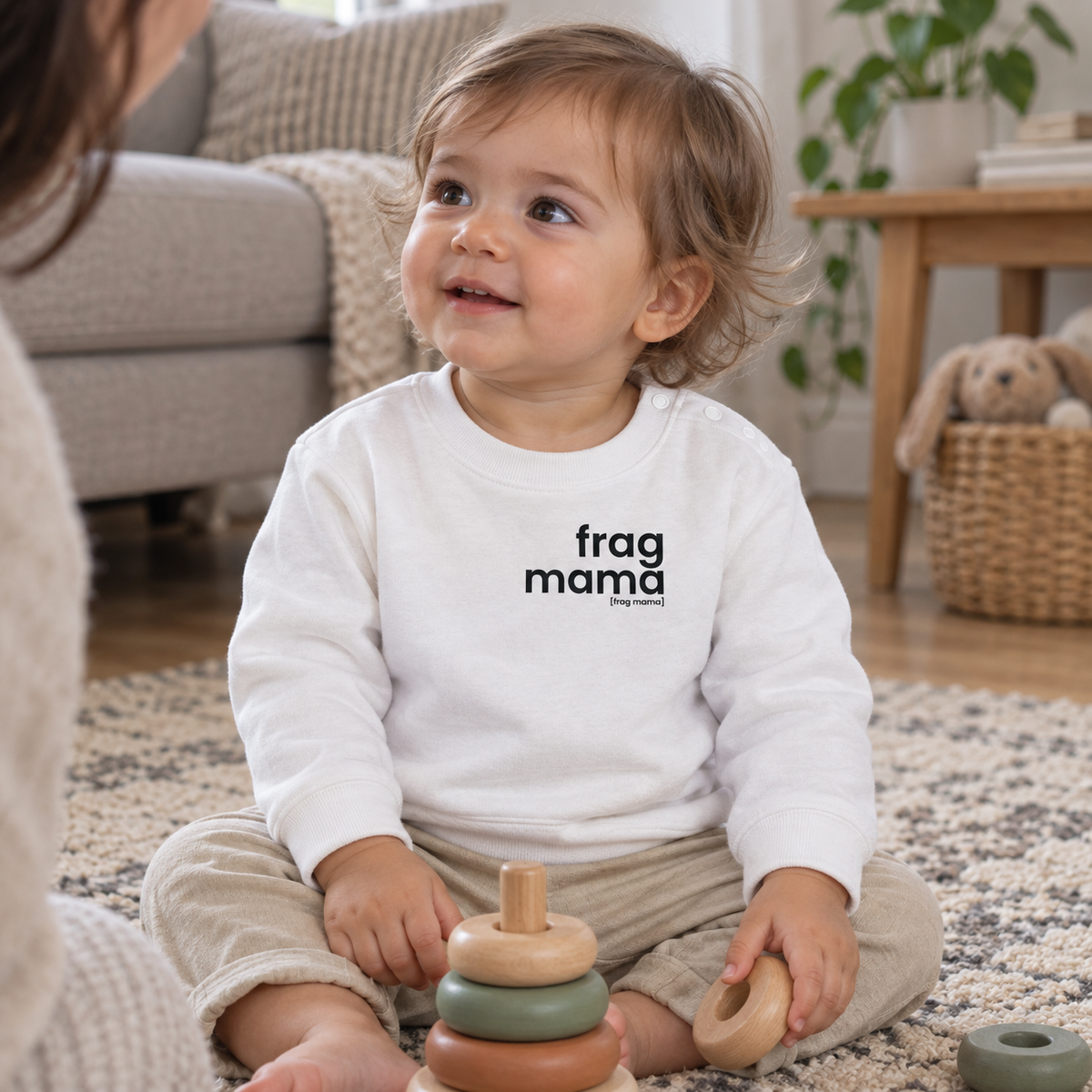 Kleinkind spielt auf dem Wohnzimmerteppich im weißen JoisPulse Baby-Sweatshirt mit dem Statement frag mama.