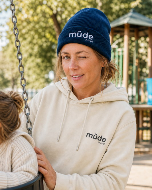 Mutter auf dem Spielplatz zwinkert, trägt die JoisPulse Beanie mit dem Statement müde