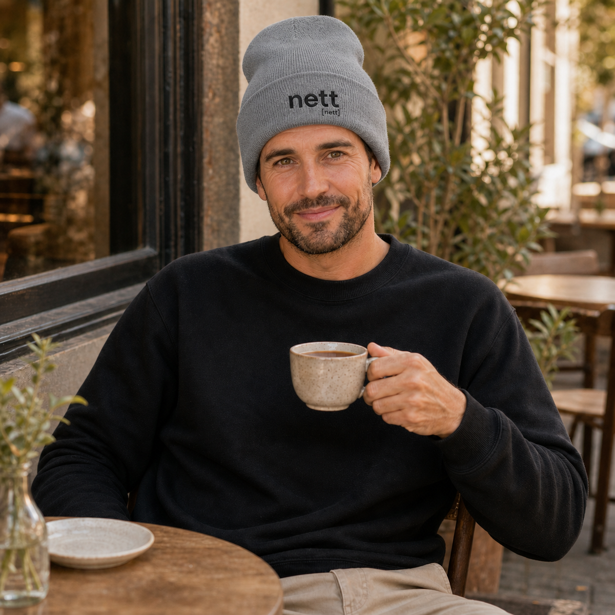 Mann im Café mit Kaffee trägt die JoisPulse Beanie mit dem Statement nett