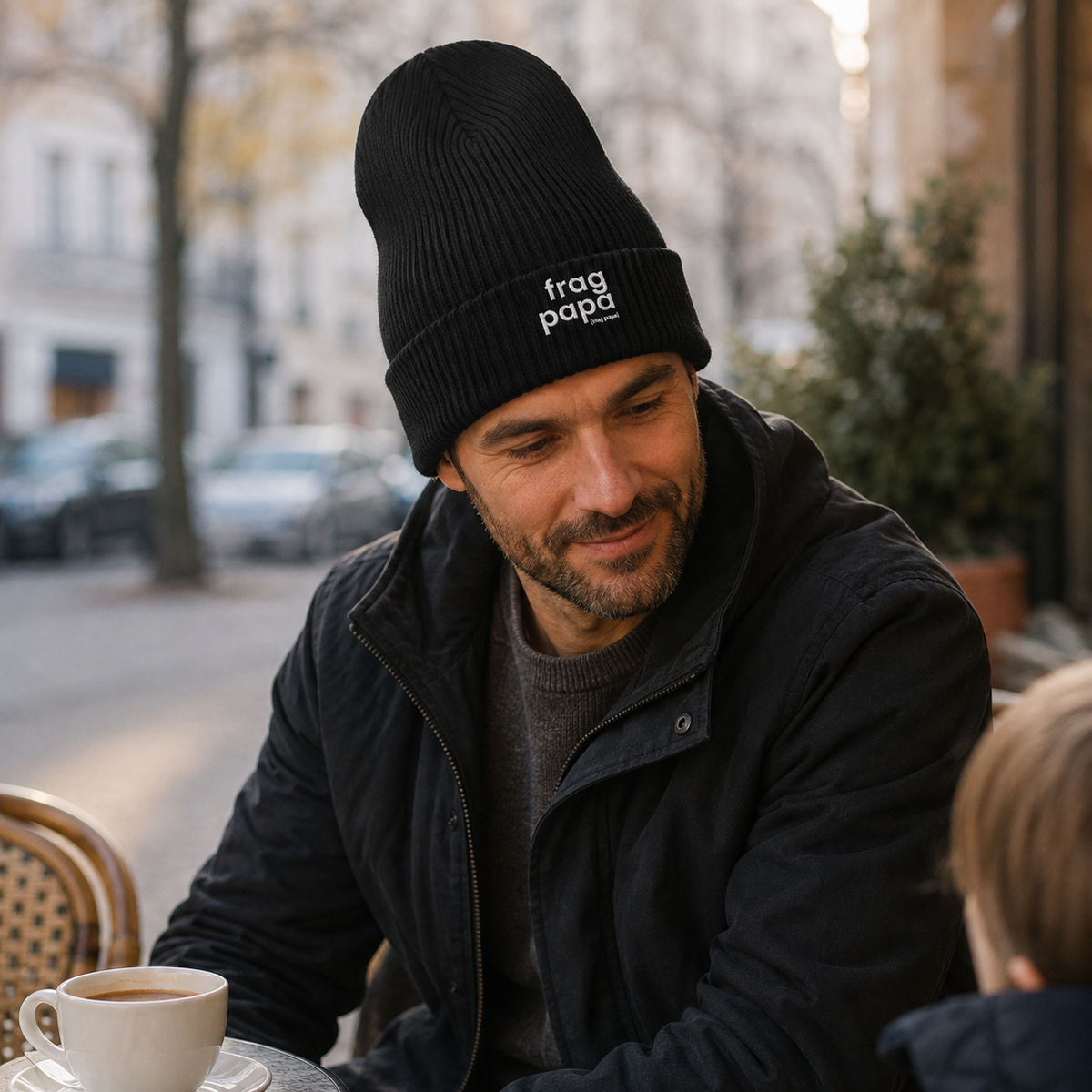 Mann im Straßencafé trägt die schwarze JoisPulse Beanie mit dem Statement frag papa