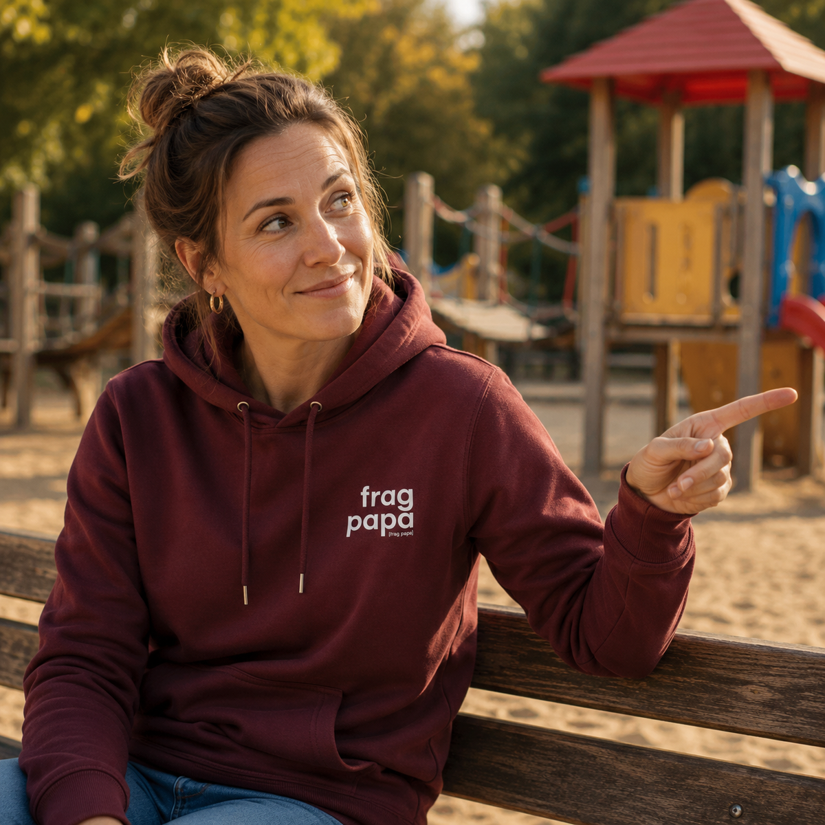 Frau auf einer Spielplatzbank im bordeauxroten JoisPulse Hoodie mit dem Statement frag papa, zeigt aus dem Bild